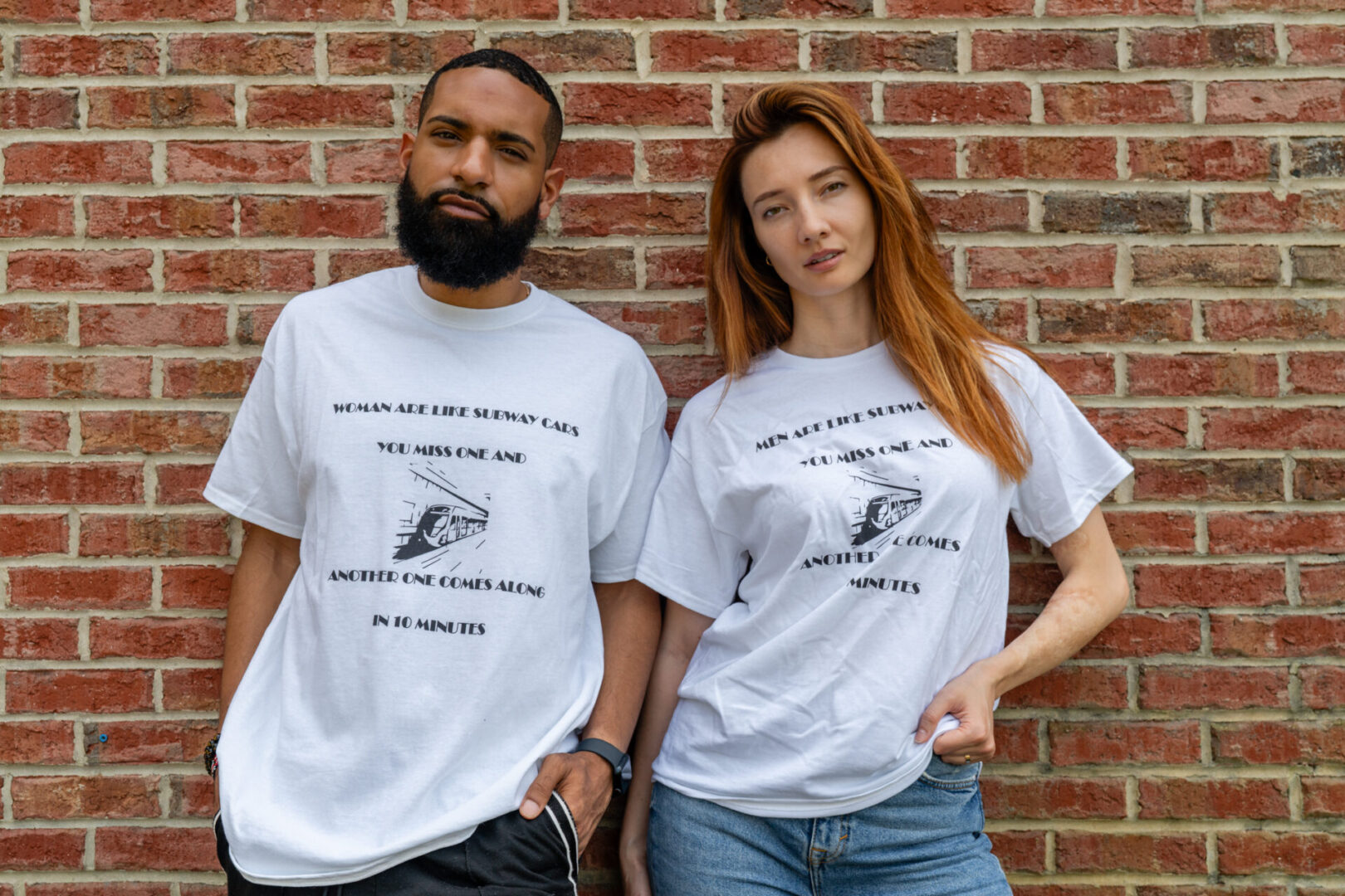 A man and woman wearing white graphic t-shirts standing against a brick wall.